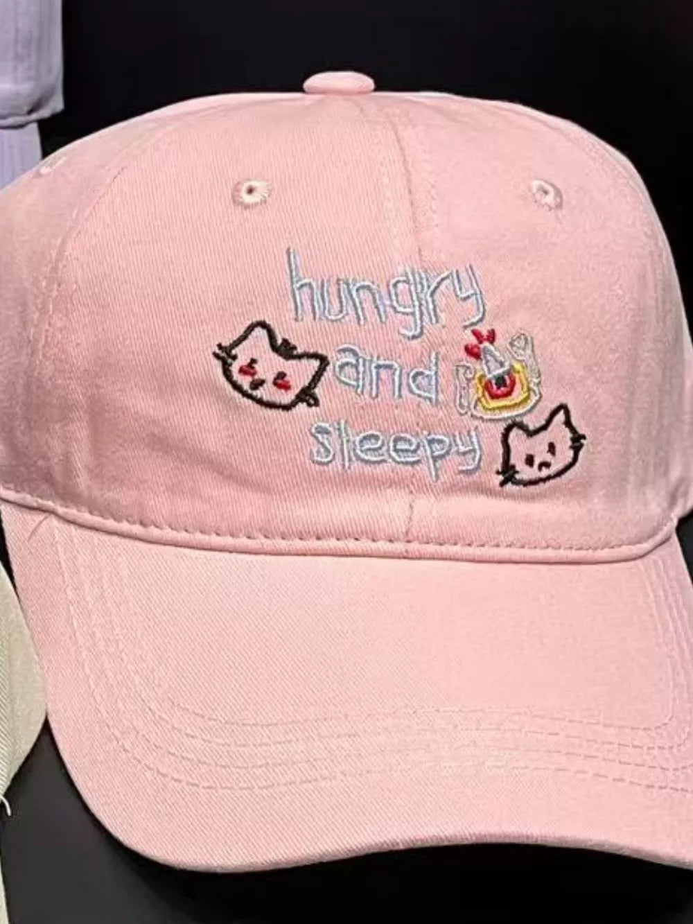 Light pink baseball cap with embroidered cats and text that reads hungry and sleepy.