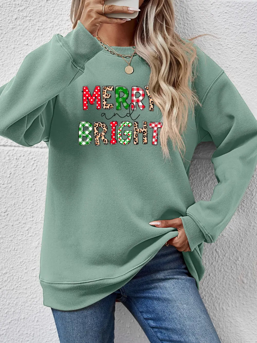 Sage green crewneck sweatshirt with "Merry and Bright" graphic in festive patterns.