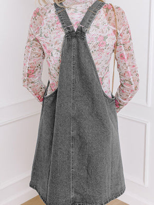 Faded gray denim pinafore dress with pockets and adjustable straps, worn over a floral top.