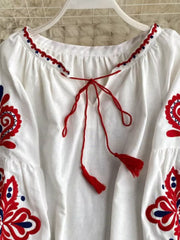 White peasant blouse with red and blue embroidery, tie neck, and tassel details.
