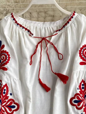 White peasant blouse with red and blue embroidery, tie neck, and tassel details.