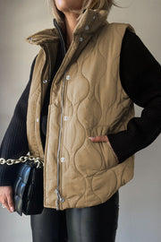 Quilted stand-collar gilet in taupe with zip and snap closure, styled with a black sweater and bag.