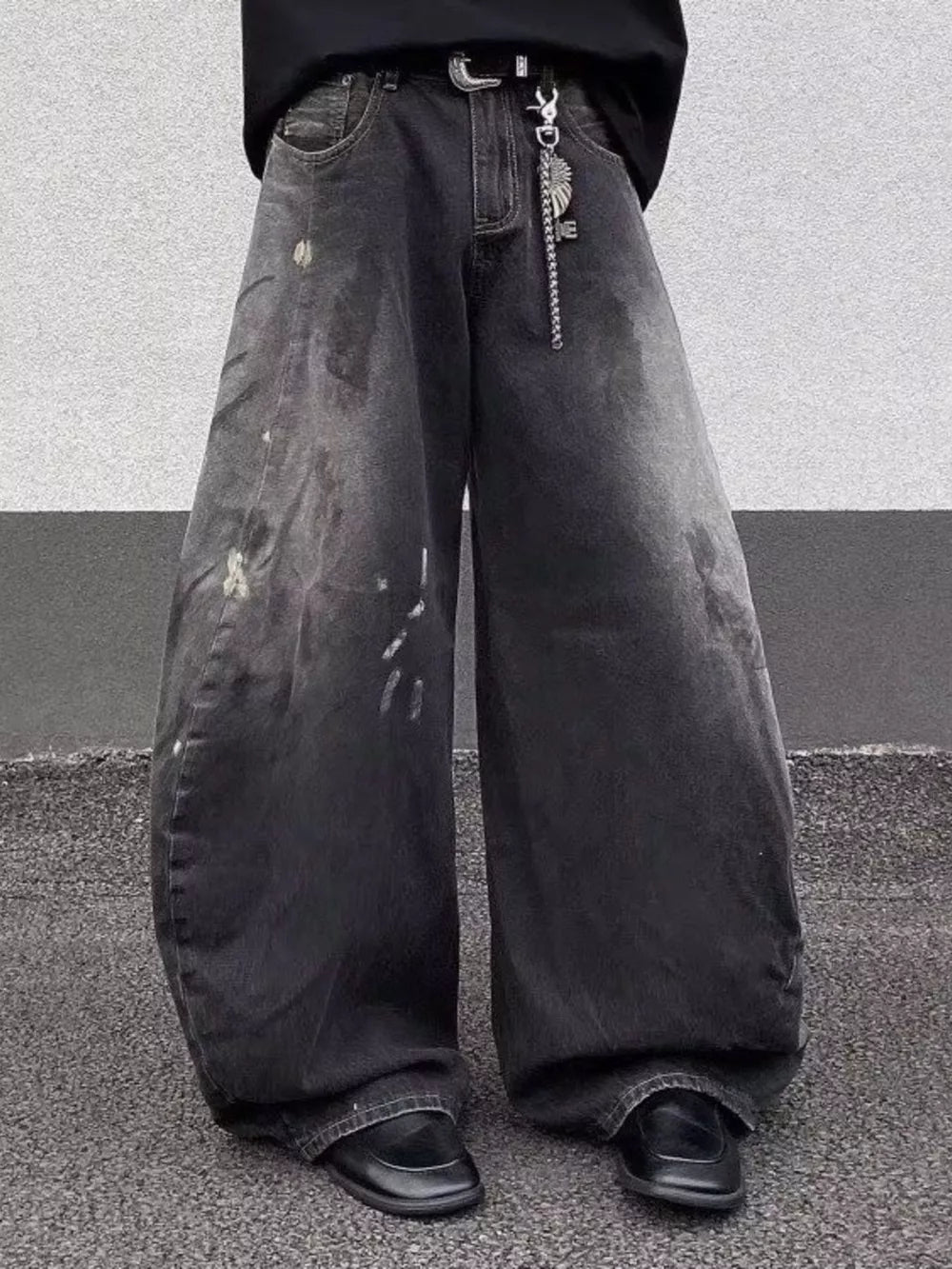 Men's black wide leg distressed jeans with paint splatters and a chain belt detail.