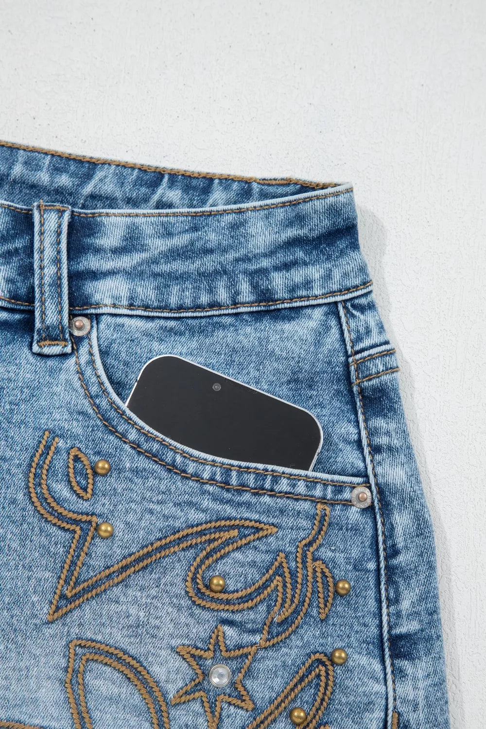 Light wash denim shorts with western embroidery, studs, and a phone in the pocket.