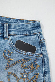 Light wash denim shorts with western embroidery, studs, and a phone in the pocket.