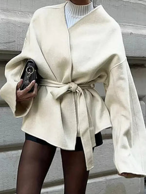 Beige belted trench coat with tie waist and dropped shoulders, styled with a black clutch.