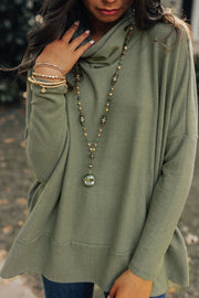 Army green tunic with cowl neck and drape front, styled with layered necklaces and bracelets.