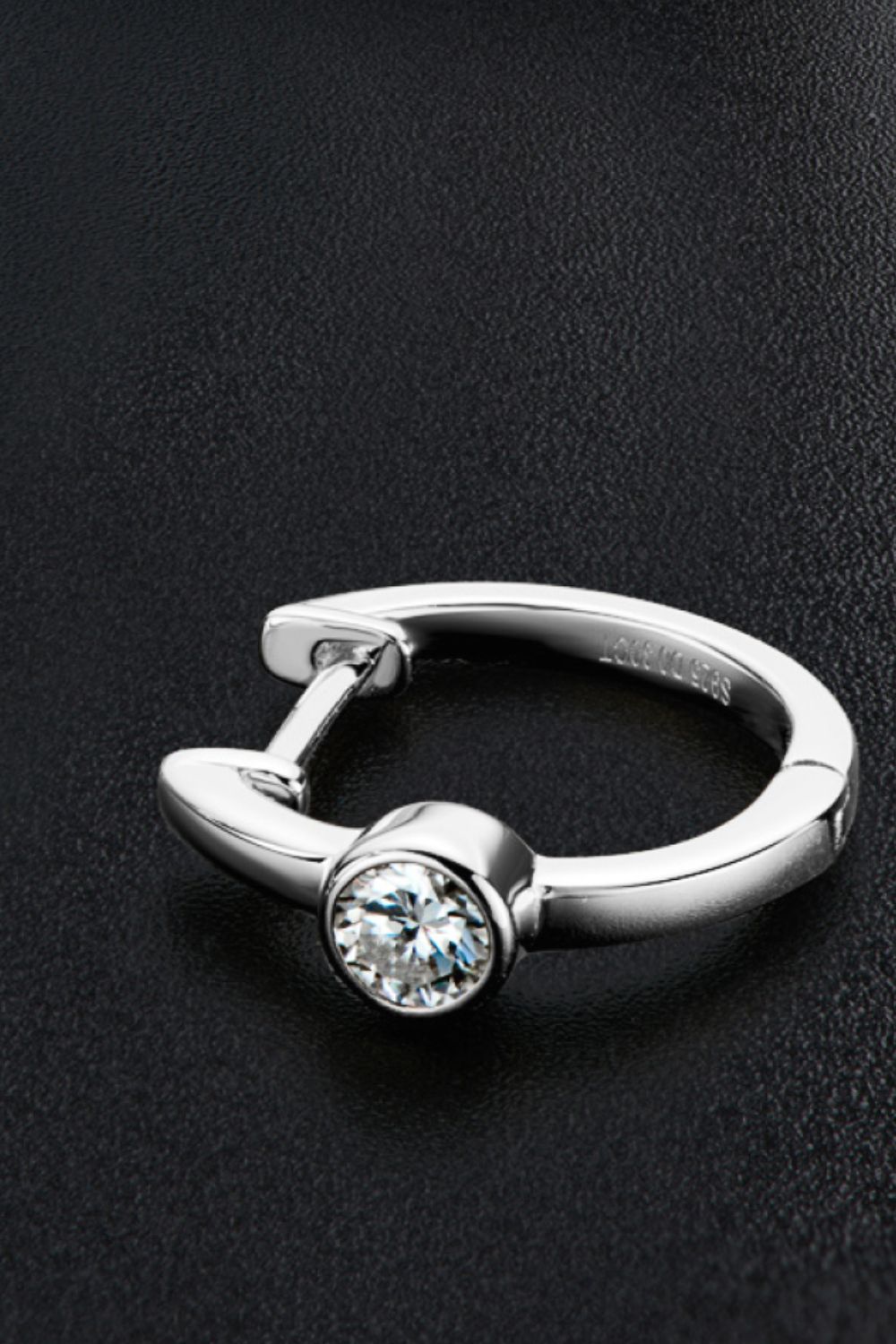 Silver huggie hoop earring with a bezel-set clear crystal on a black textured background.