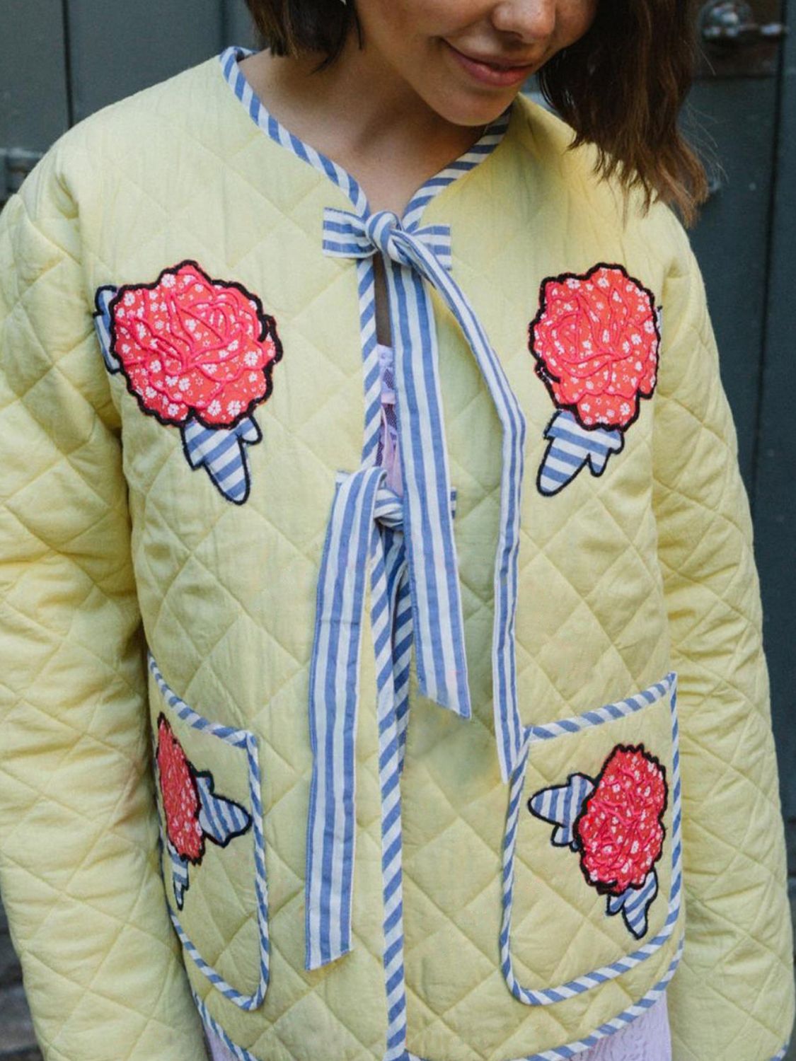 Pastel yellow quilted jacket with floral appliqué, blue striped trim, and tie closure.