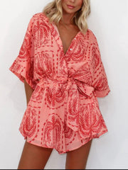 Coral wrap romper with red palm tree print, short sleeves, and tie waist.