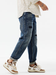 Men's dark blue distressed denim jeans with patchwork details and cuffed hems.