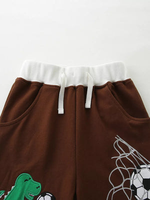 Brown cotton baby shorts with drawstring, white waistband, and dinosaur and soccer graphics.