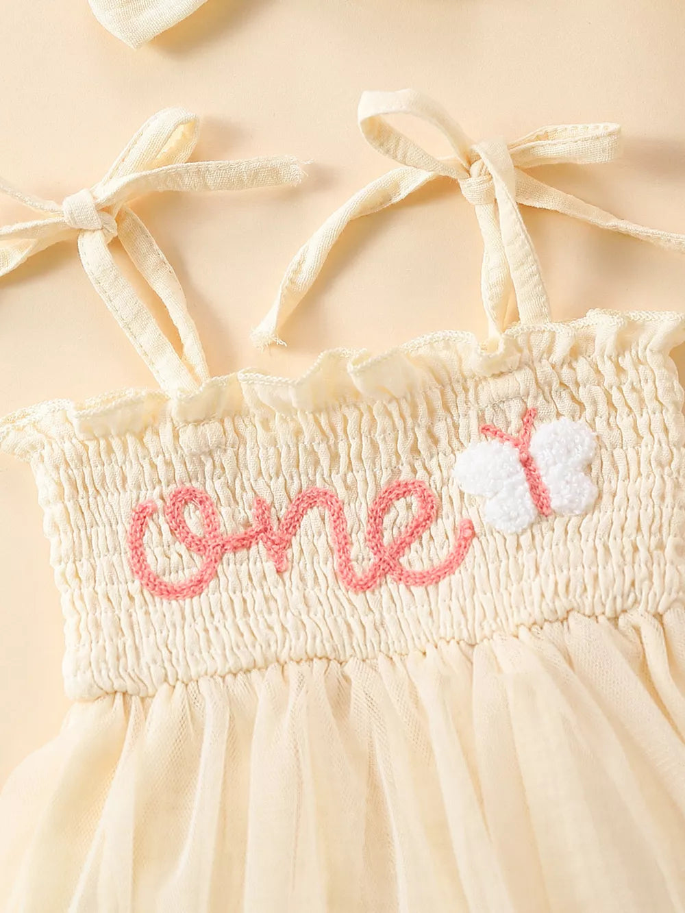 Cream smocked bodysuit with pink "one" embroidery and butterfly detail, with tie straps.