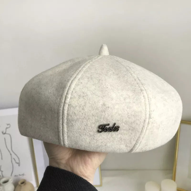 Ivory woolen beret with distinct paneled design and metal logo detail.
