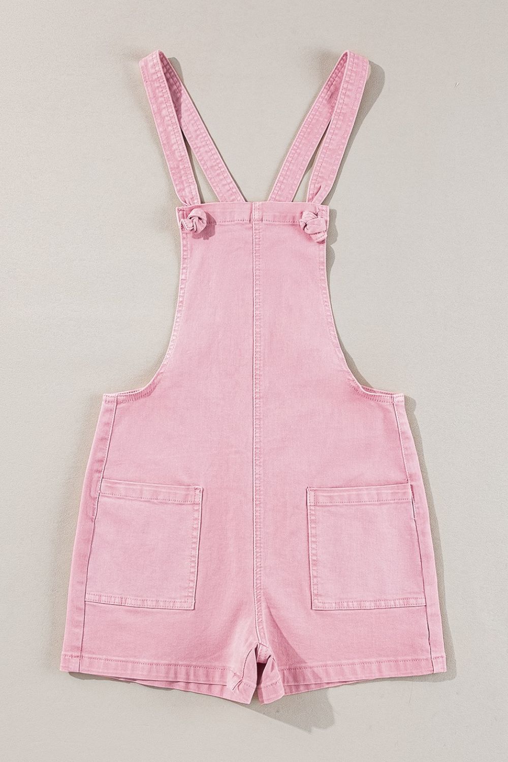 Blush pink denim short overalls with knotted straps and patch pockets.
