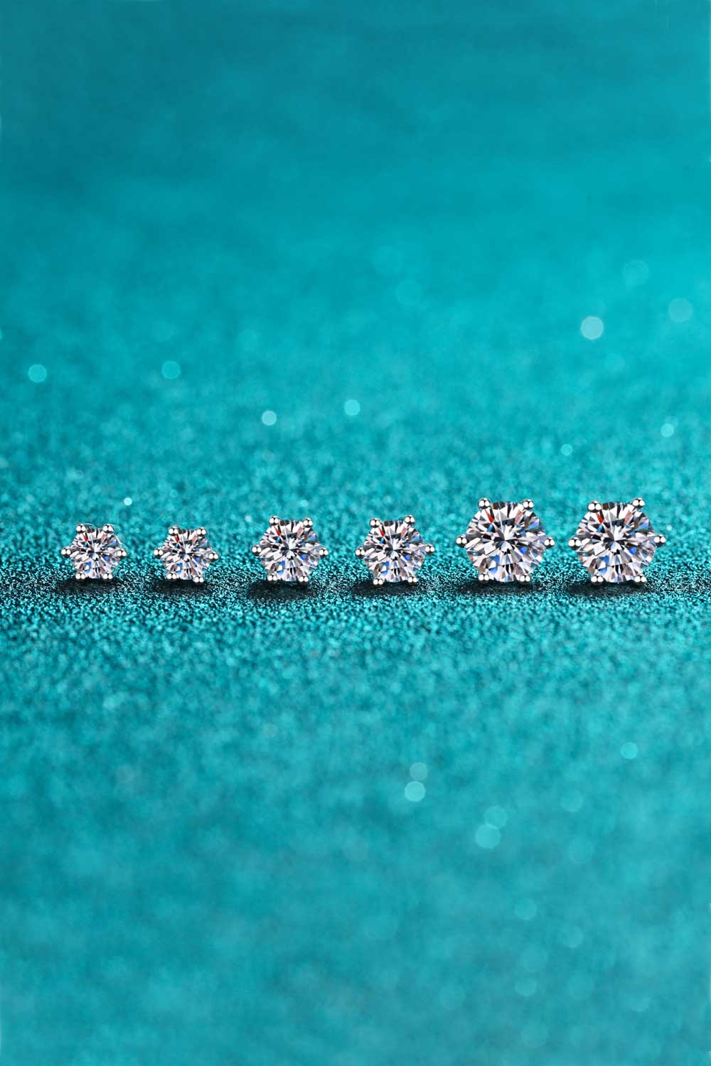 Six-prong moissanite solitaire stud earrings in silver, showcasing different sizes on a teal glitter background.