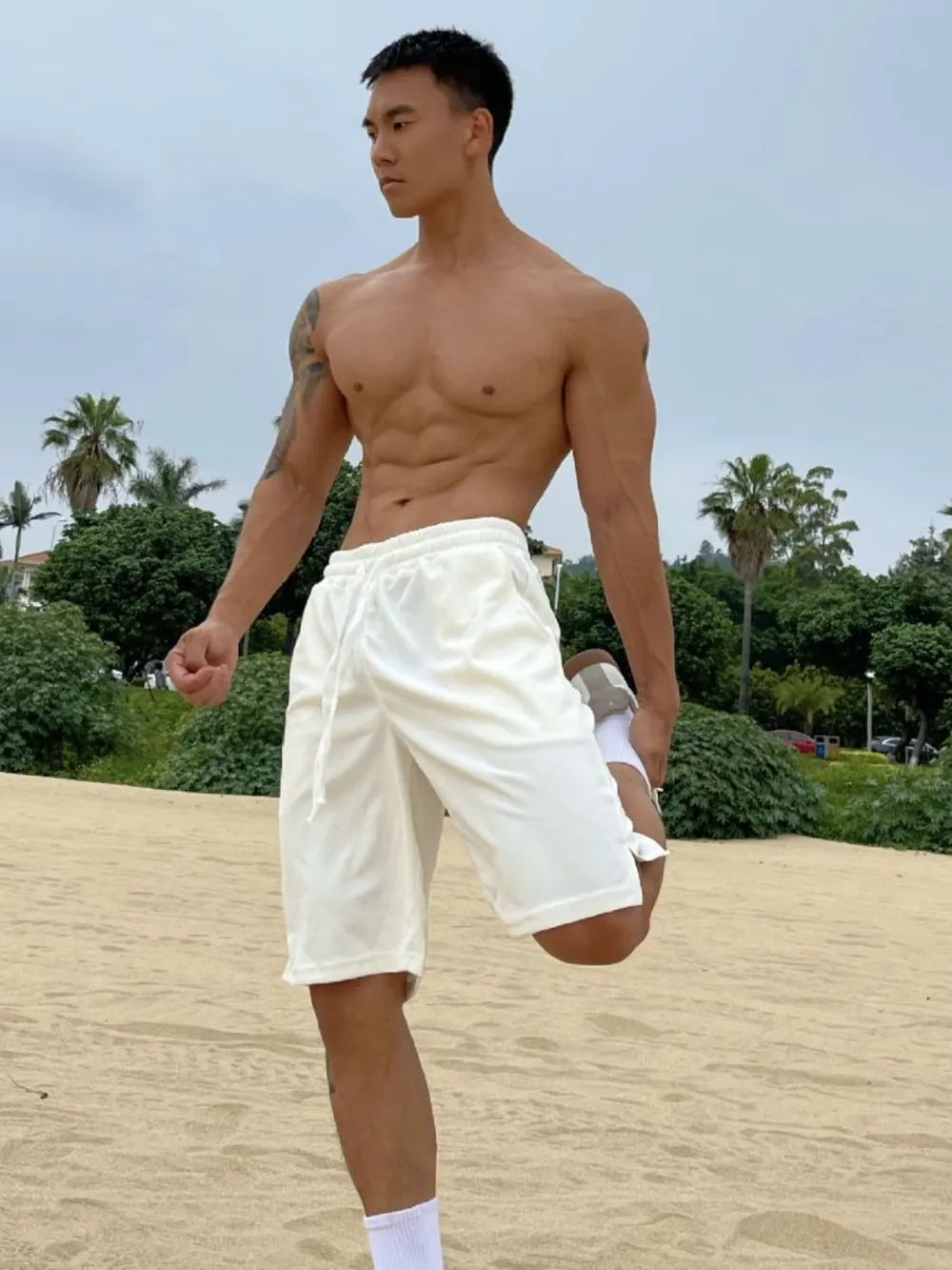 Relaxed-fit white drawstring bermuda shorts with pockets, on a man stretching on the beach.