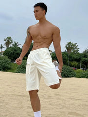Relaxed-fit white drawstring bermuda shorts with pockets, on a man stretching on the beach.