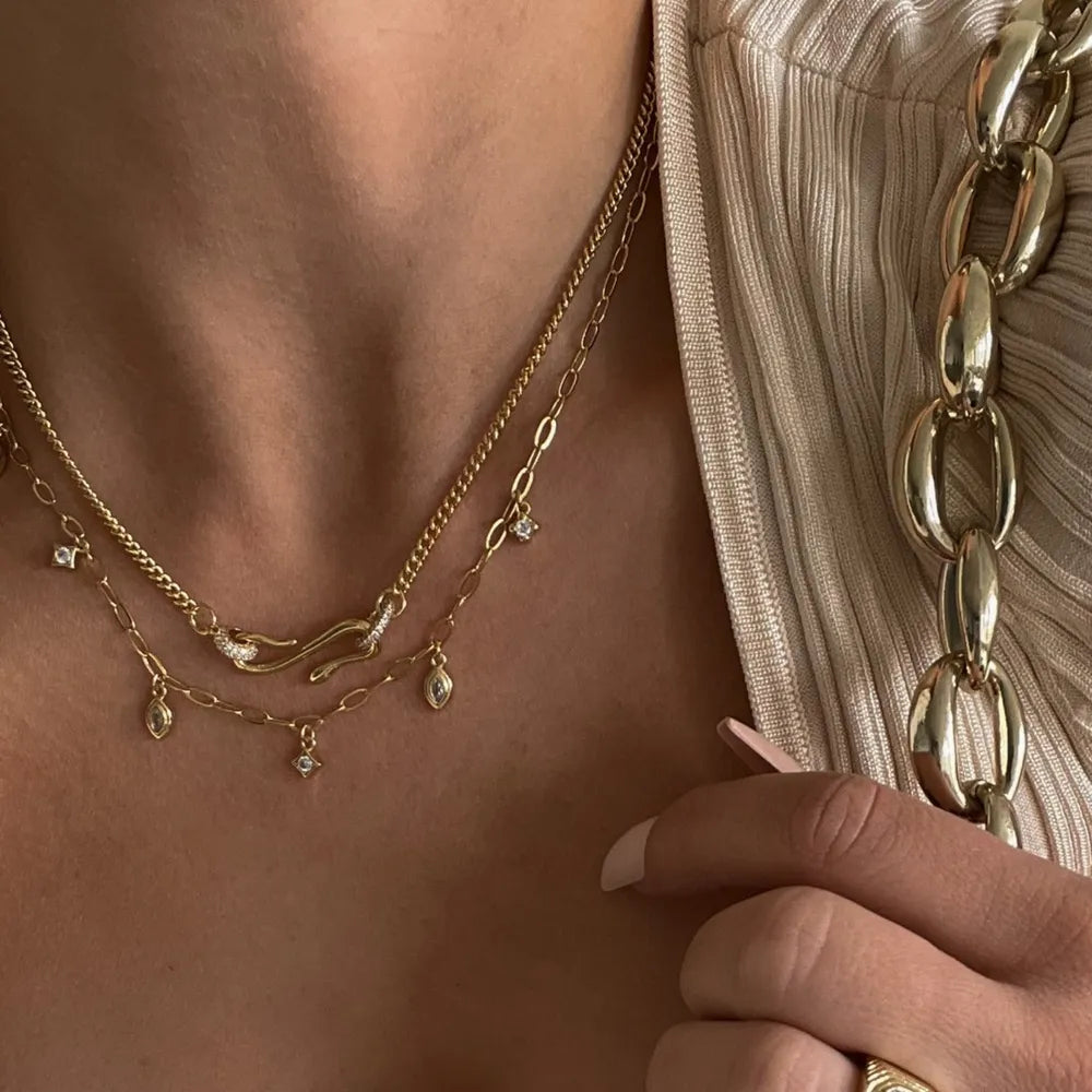 Gold necklaces with cubic zirconia charms and a serpentine pendant layered on a beige top.
