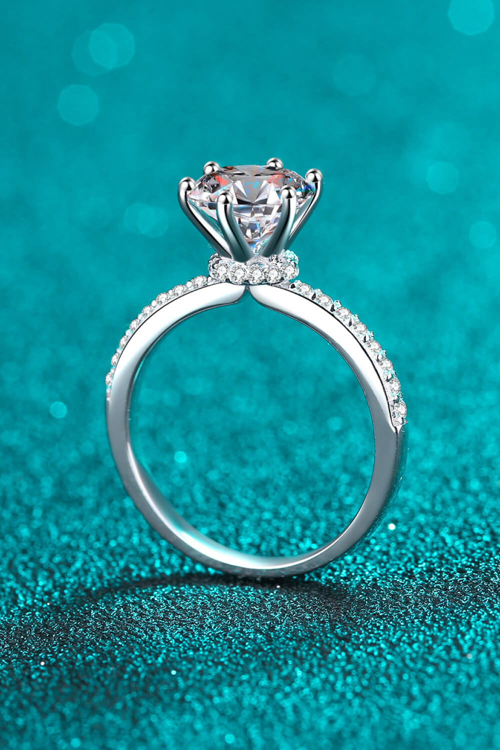 Silver moissanite ring with pave band and solitaire diamond on a teal glitter background.