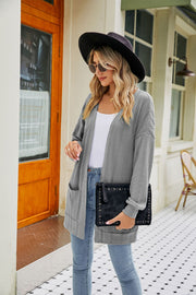 Chic grey cable-knit cardigan with pockets and a relaxed fit, styled with jeans and a clutch.