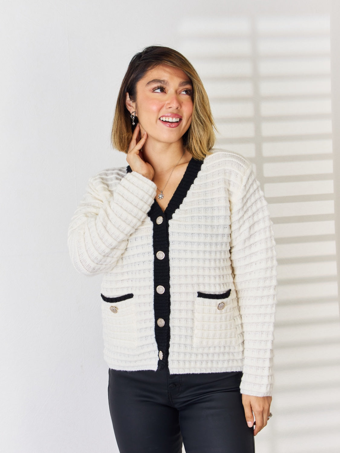 White textured knit cardigan with black contrast trim, buttons, and pocket details.
