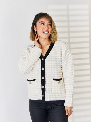 White textured knit cardigan with black contrast trim, buttons, and pocket details.