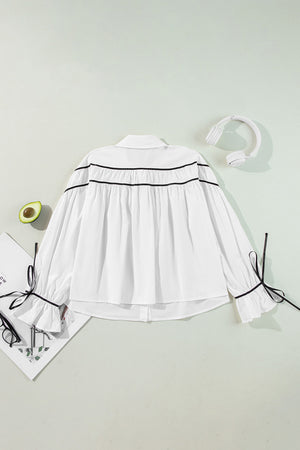 White blouse with black piped trim and bow details on ruffled cuffs.