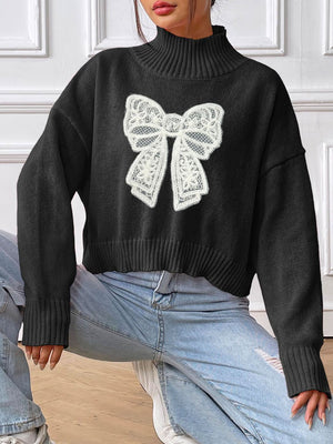 Black turtleneck knit sweater with white lace bow appliqué detail.
