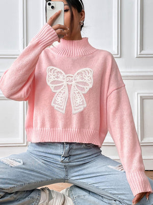 Pink turtleneck knit sweater with white lace bow appliqué and ribbed cuffs and hem.