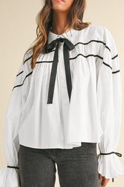 White blouse with black piped trim and bow detail, featuring ruffled cuffs.