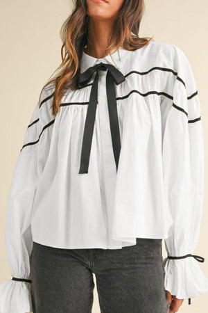 White blouse with black piped trim and bow detail, featuring ruffled cuffs.