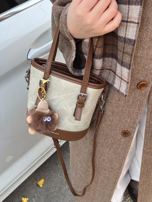 Premium two-tone bucket crossbody bag in beige canvas with brown trim and fuzzy charm.