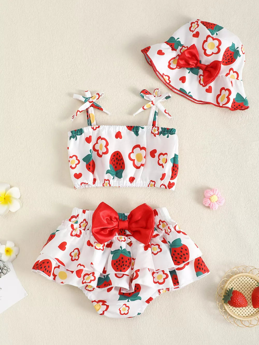 Baby strawberry print set with ruffled bloomers, tie-strap top, and bucket hat with red bow.