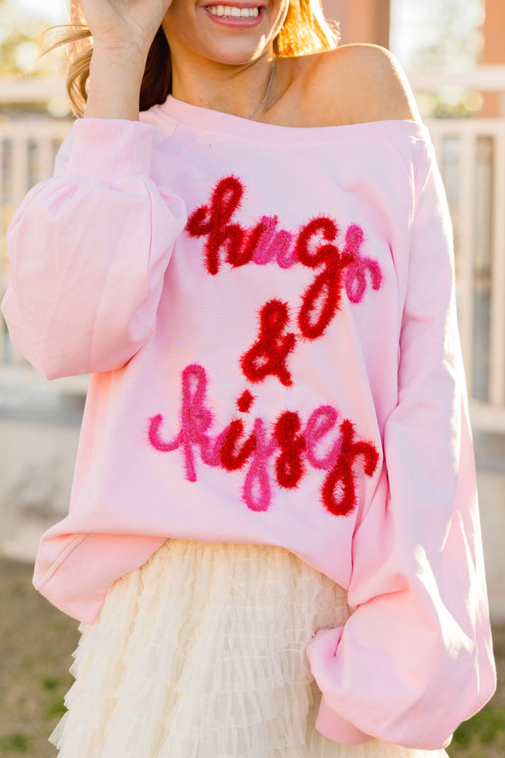 Off-shoulder blush pink sweatshirt with fuzzy red and pink "hugs & kisses" graphic and balloon sleeves.