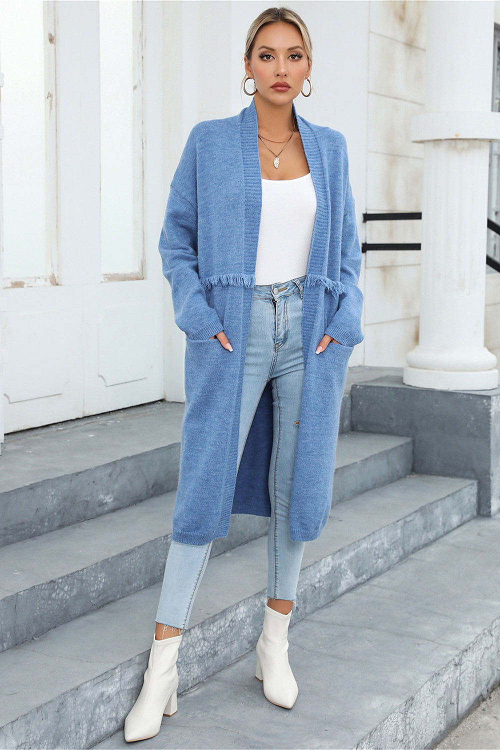 Light blue longline knit cardigan with fringe trim and pockets, worn open over a white top and jeans.