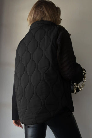 Black quilted stand-collar utility gilet with a wave pattern, worn over a black sweater.
