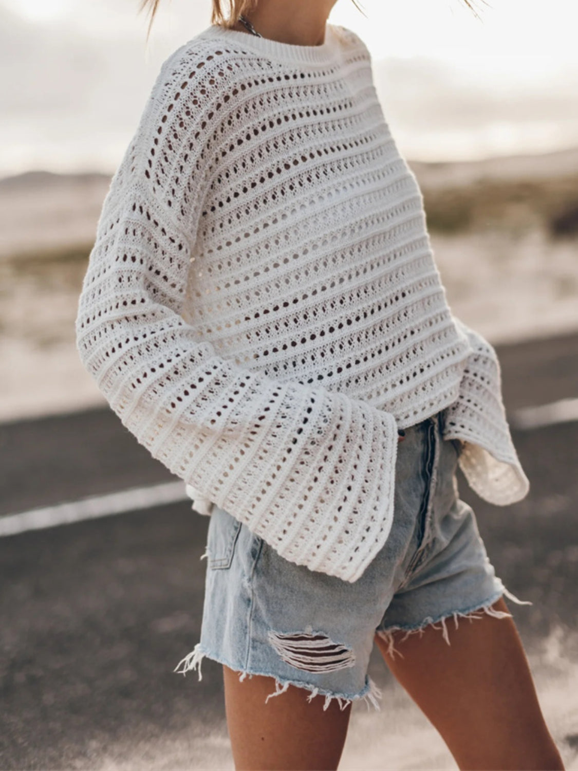 White open-knit sweater with bell sleeves and a crew neck, styled with denim shorts.