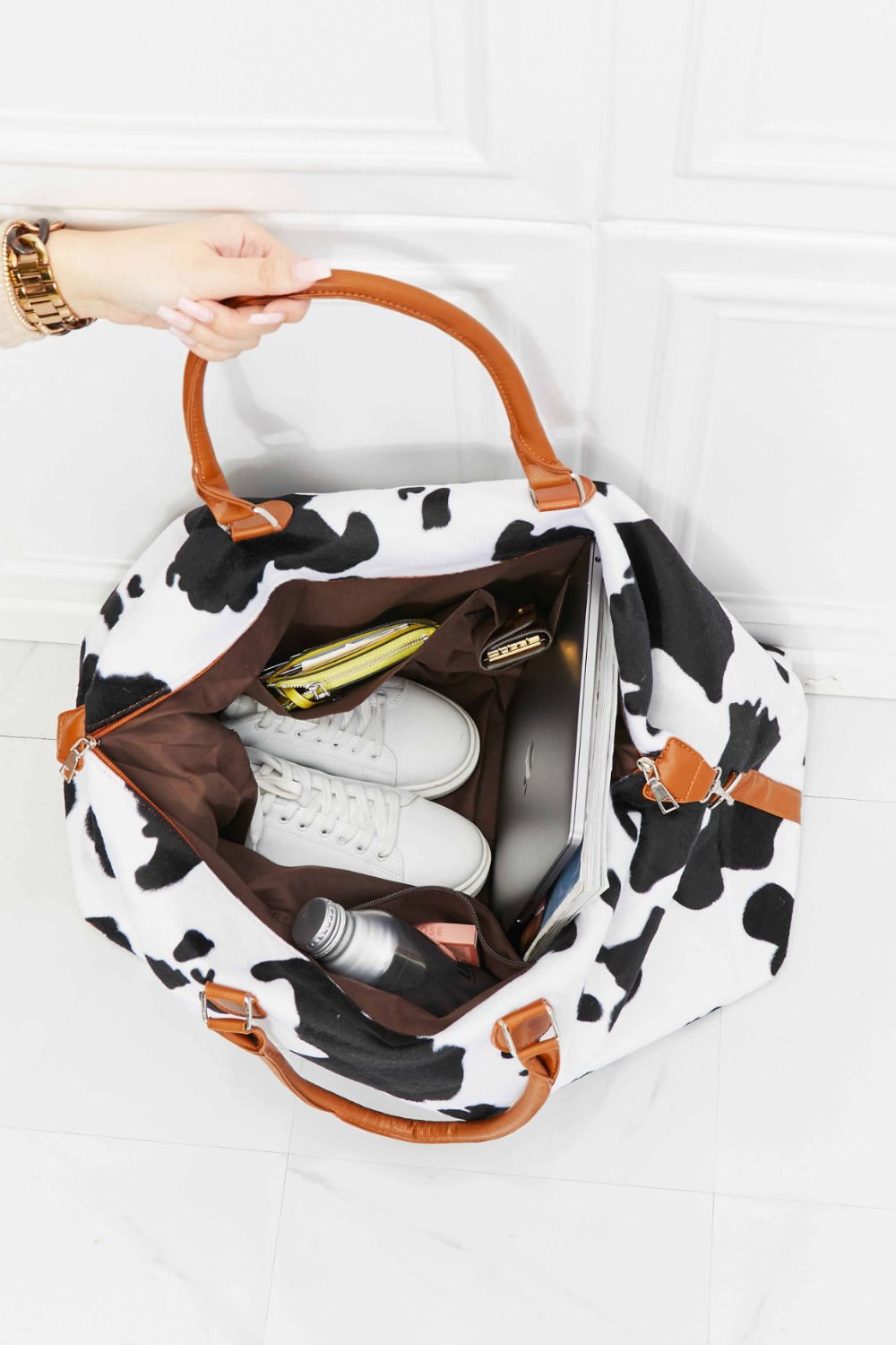 Cowhide print duffel bag with brown leather handles, packed with shoes, laptop, and accessories.