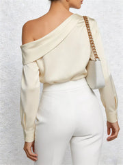 Cream one-shoulder draped blouse with long sleeves, styled with white pants and a chain bag.