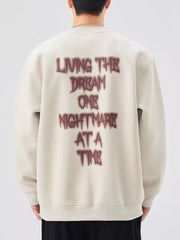 Oversized crew-neck sweatshirt in light beige with dark red "Living the dream one nightmare at a time" graphic on back.