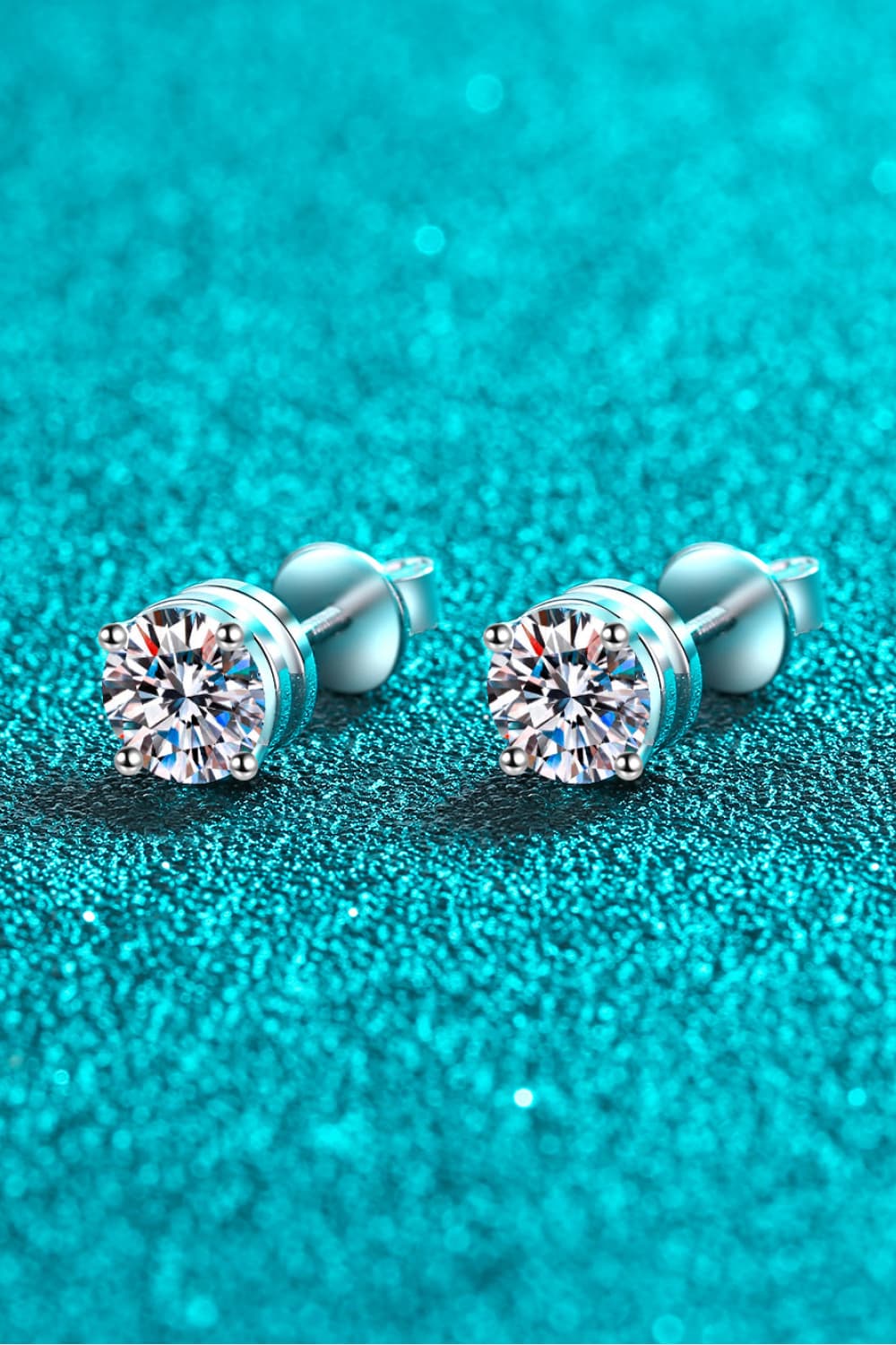 Moissanite solitaire stud earrings with rhodium finish on a teal glitter background.