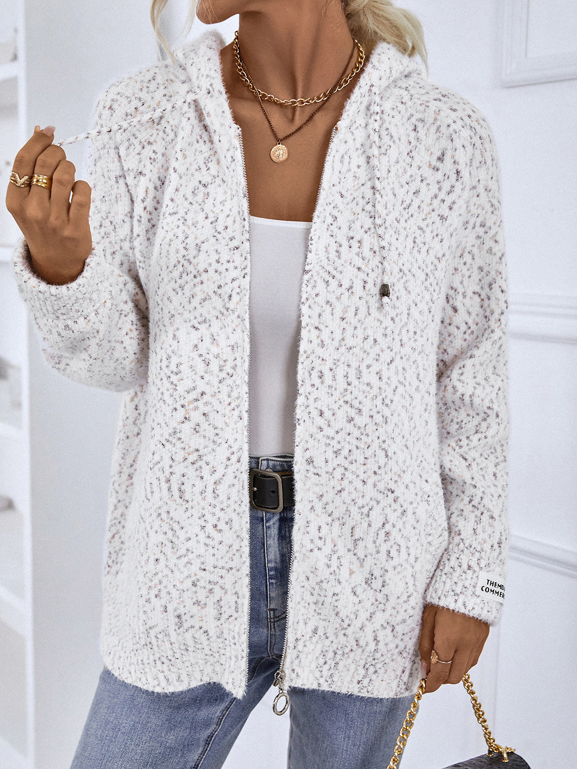 Textured knit zip-up cardigan in white with speckled pattern, drawstring hood, and logo detail on sleeve.