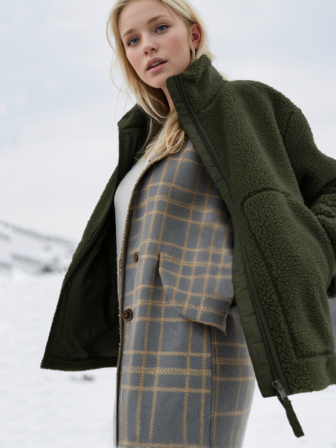 Olive green sherpa jacket with stand collar and zipper detail, worn over a plaid vest.