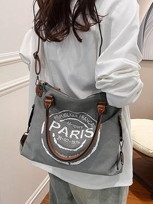 Parisian graphic tote bag in dark gray canvas with brown handles and airport stamp detail.