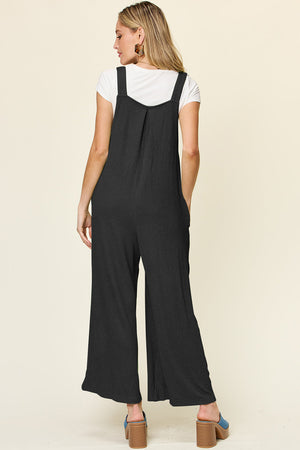 Black ribbed wide-leg pinafore jumpsuit with pockets and a pleated bodice detail.