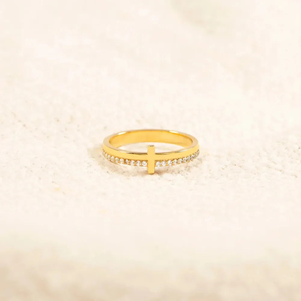 Gold pavé cross band ring with clear stones on a textured white surface.