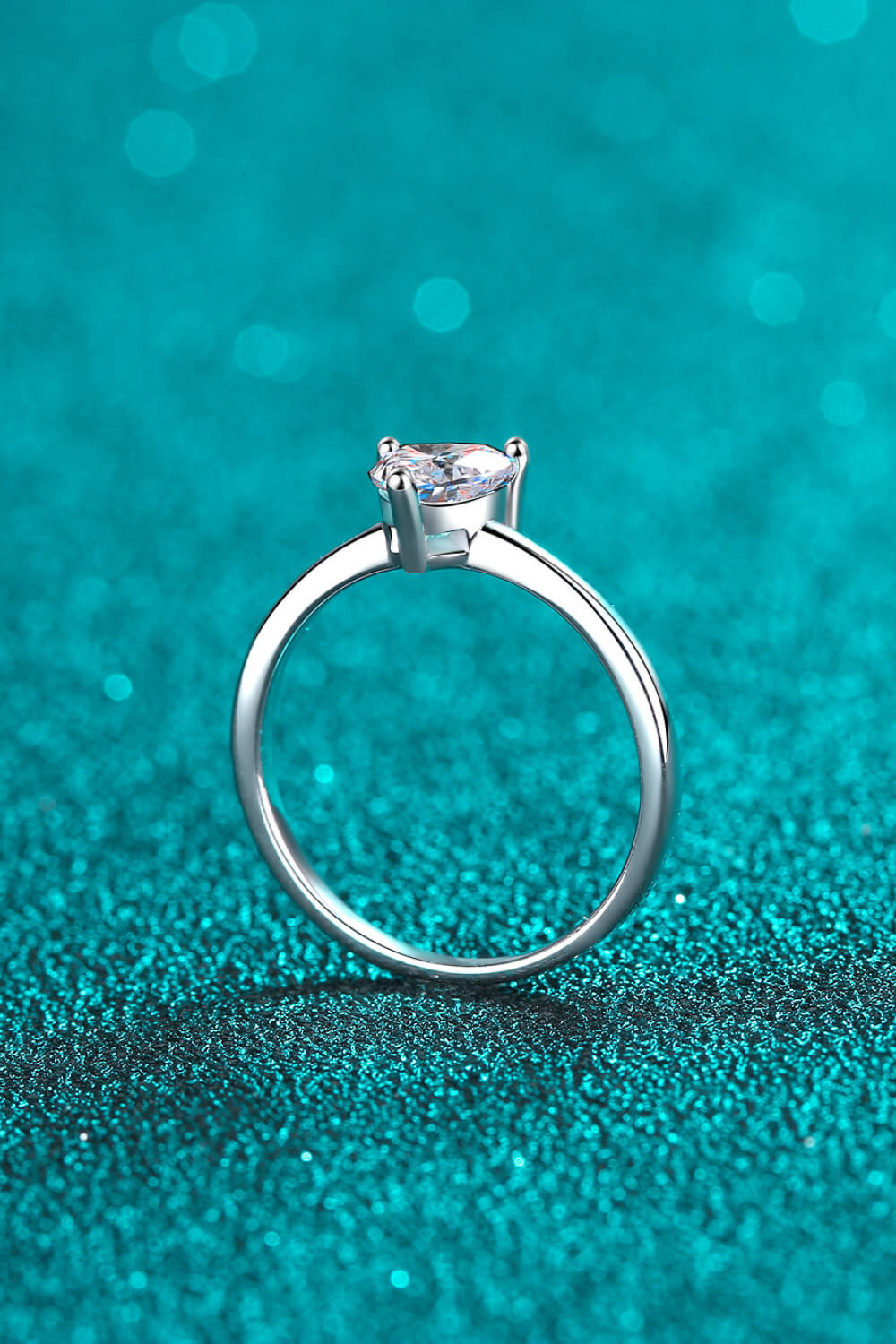 Silver heart-cut moissanite solitaire ring on a sparkly teal background.