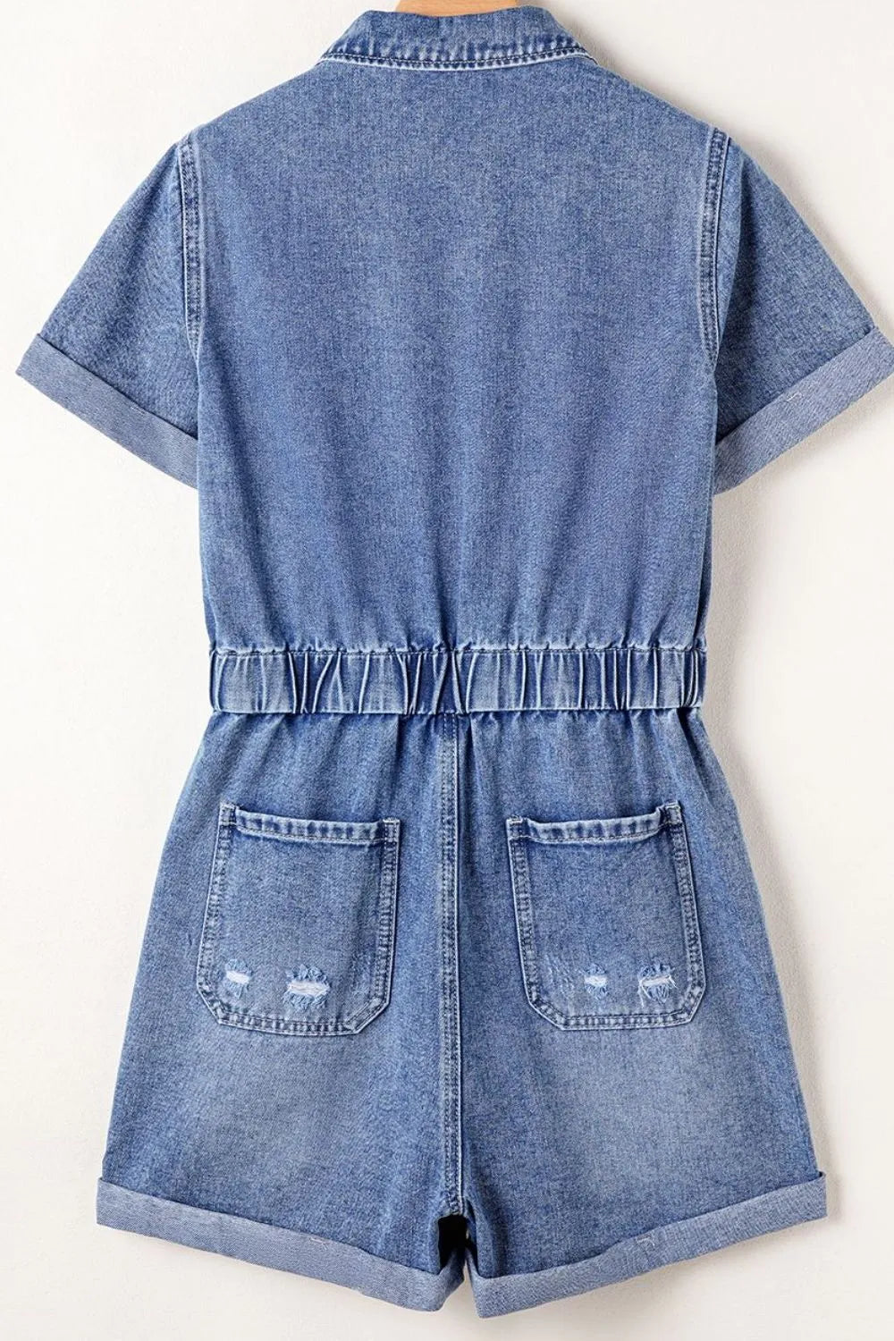 Medium wash denim romper with elastic waist, short sleeves, and distressed back pockets.