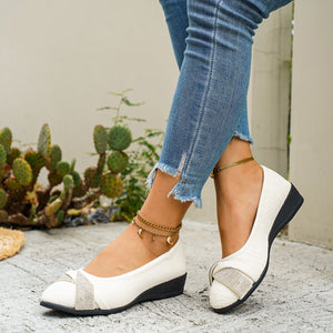 Beige slip-on wedge flats with rhinestone bow detail and textured fabric.